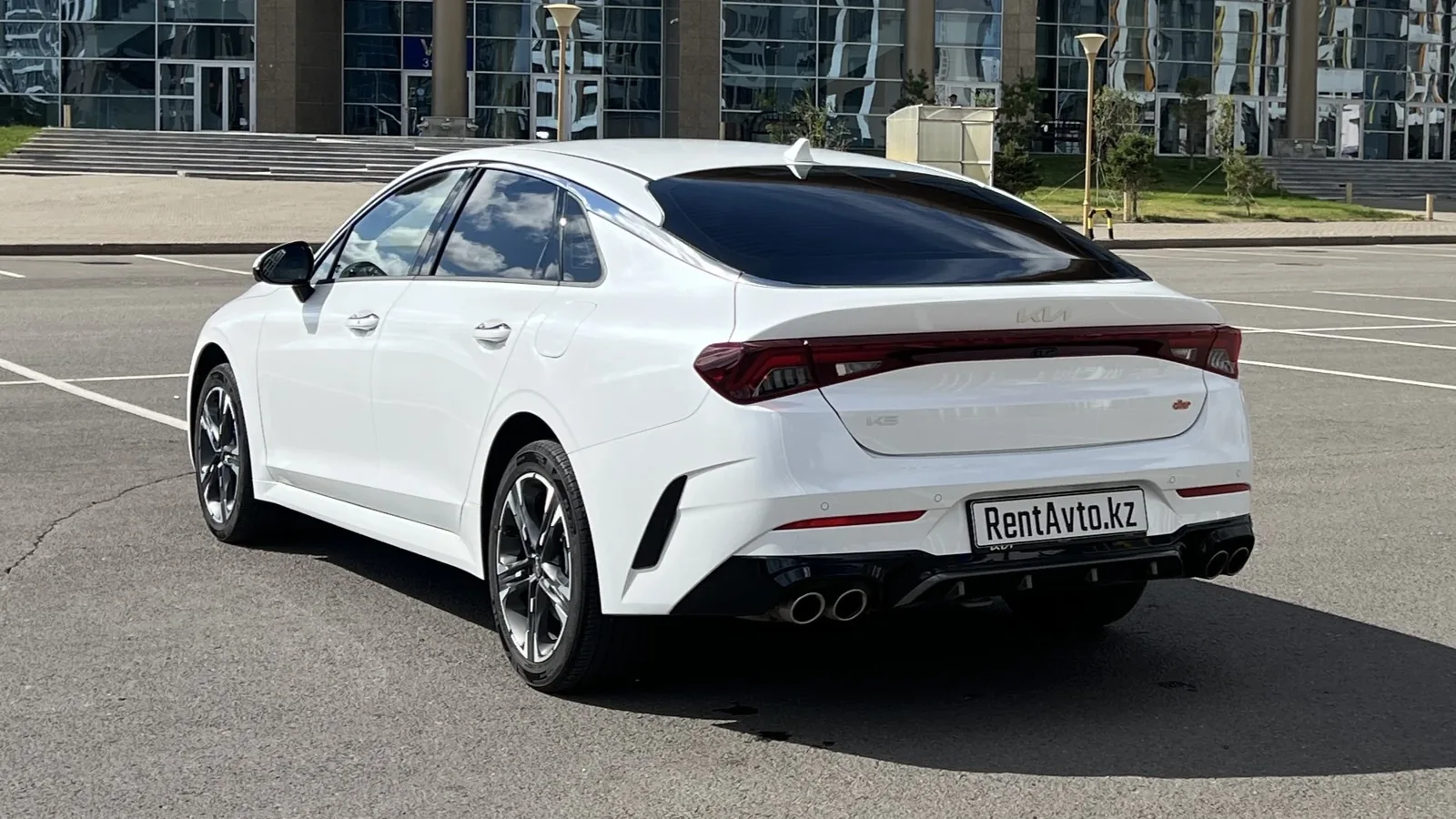 Белая KIA K5 снята сзади на фоне деловых зданий, легковой бизнес-седан подготовлен к подаче с водителем