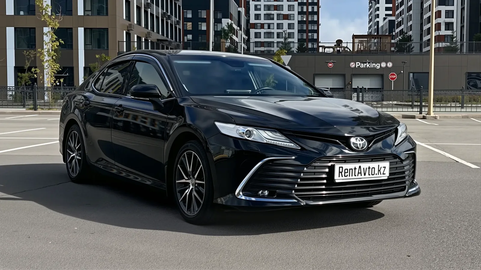 Чёрная Toyota Camry 75 снята спереди на фоне деловых зданий, седан подают с водителем для поездок по городу