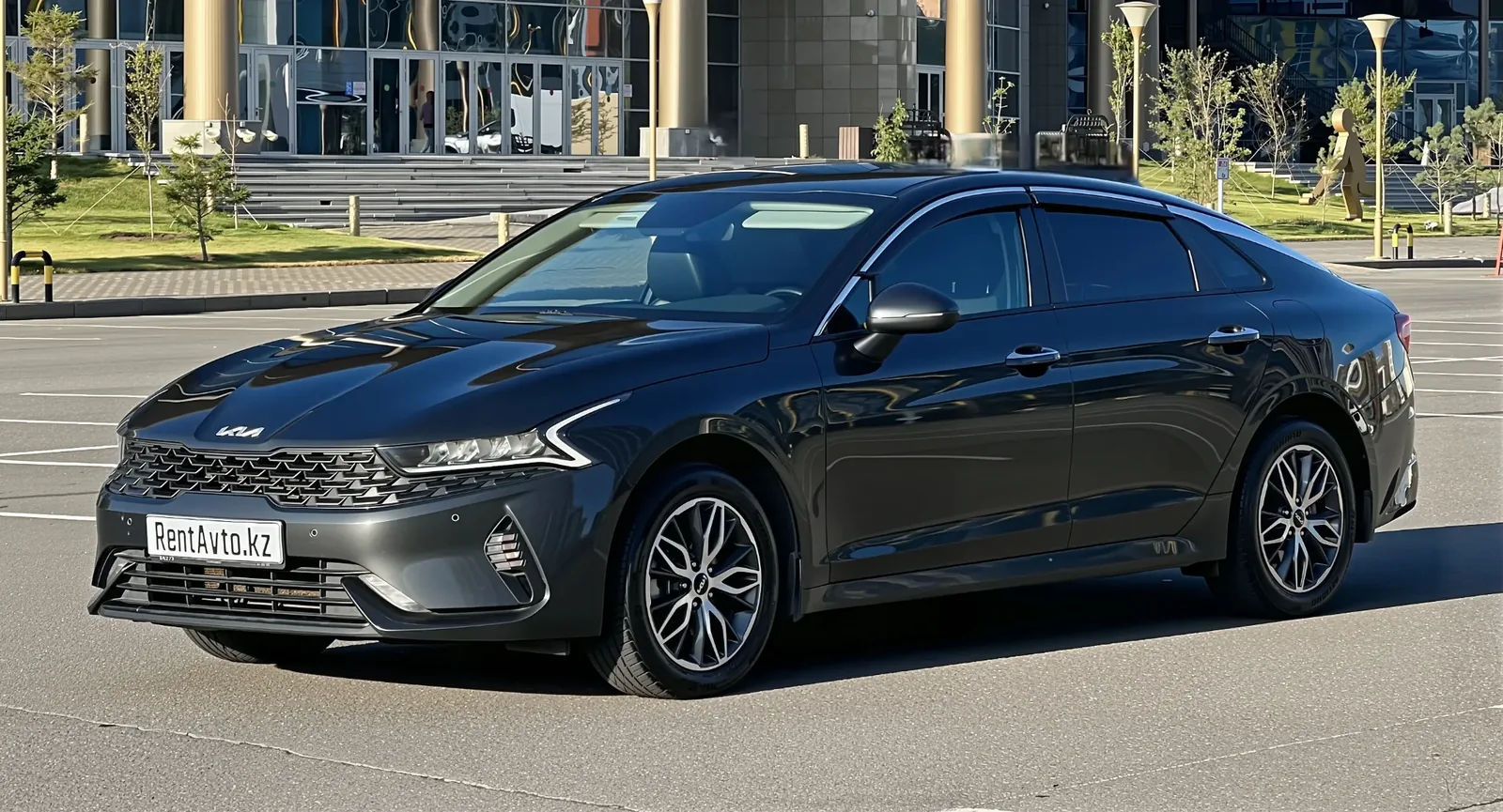 KIA K5 сфотографирована спереди на открытой площадке, автомобиль используют для поездок с водителем из Астаны в Боровое