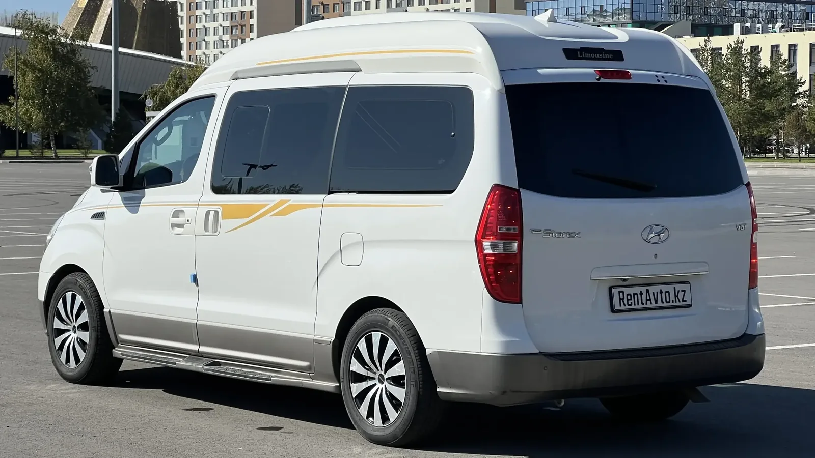 Белый Hyundai Grand Starex показан сбоку на ровной асфальтовой площадке с дорожной разметкой, находящийся на прокате у клиентов
